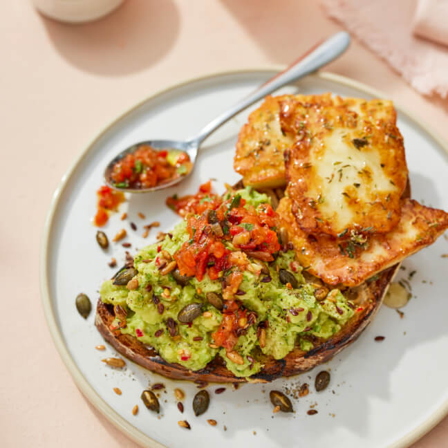 Avocado toast with mashed avocado, red pepper salsa, mixed seeds, and grilled halloumi on a white plate.