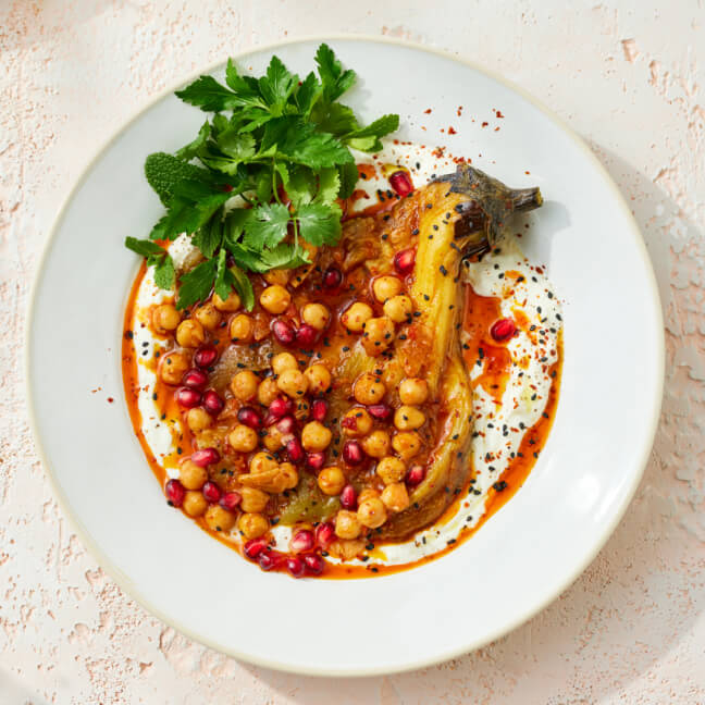 Roasted eggplant served on a bed of yogurt, topped with spiced chickpeas, pomegranate seeds, and fresh herbs, garnished with chili oil and black sesame seeds on a white plate.