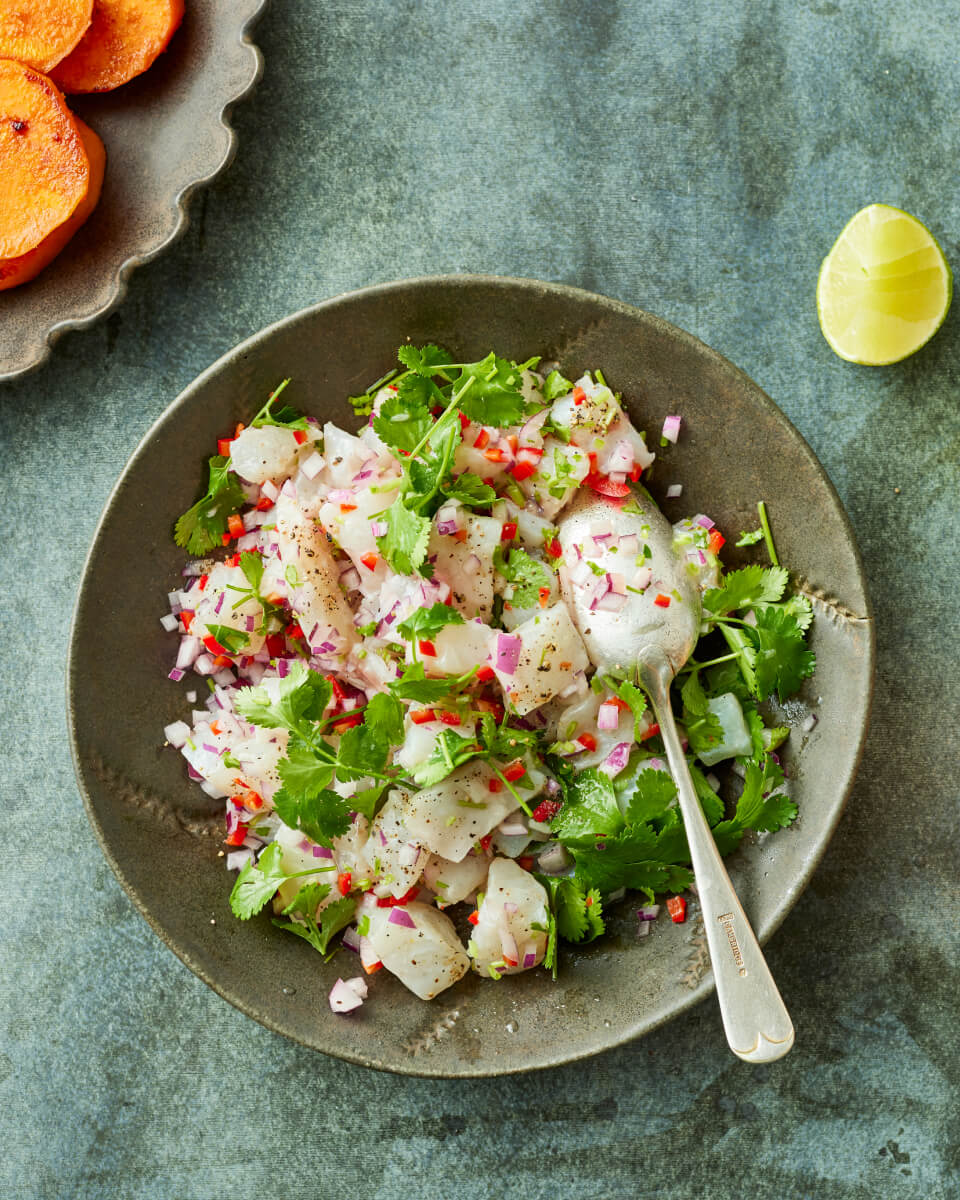 A plate of fresh fish ceviche garnished with chopped red onion, chili, cilantro, and black pepper, served with a spoon on a rustic surface, with a lime wedge and sliced sweet potatoes on the side.