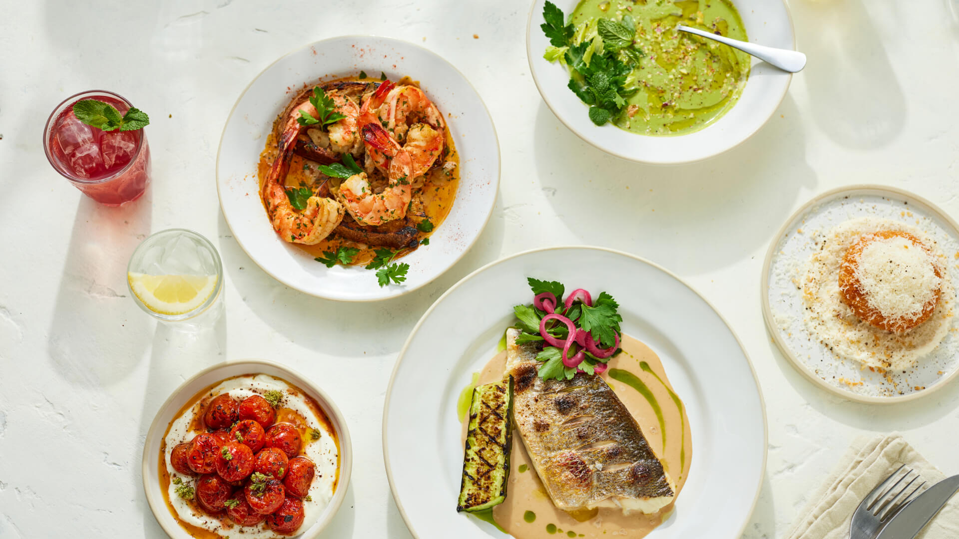 A spread of dishes including shrimp in sauce, green soup, grilled fish with garnish, cherry tomatoes with herbs, dessert, and drinks on a white table.