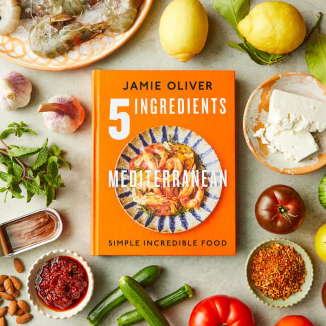 Jamie Oliver's cookbook titled “5 Ingredients Mediterranean: Simple Incredible Food” placed on a countertop surrounded by fresh ingredients including prawns, lemons, feta cheese, garlic, tomatoes, zucchini, mint, chili paste, almonds, and spices.