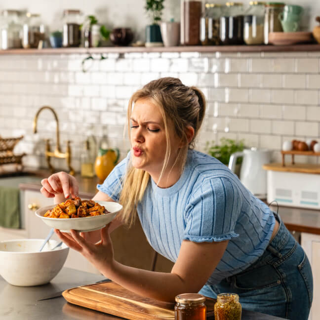 Woman in a blue ribbed top holding a bowl of patatas bravas and making an expressive face in a modern kitchen, with jars, plants, and utensils on the counter and shelves in the background.