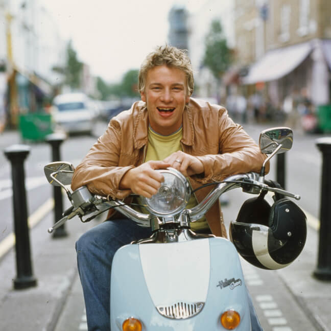 A young Jamie Oliver on a light blue vintage scooter, wearing a brown leather jacket and smiling.